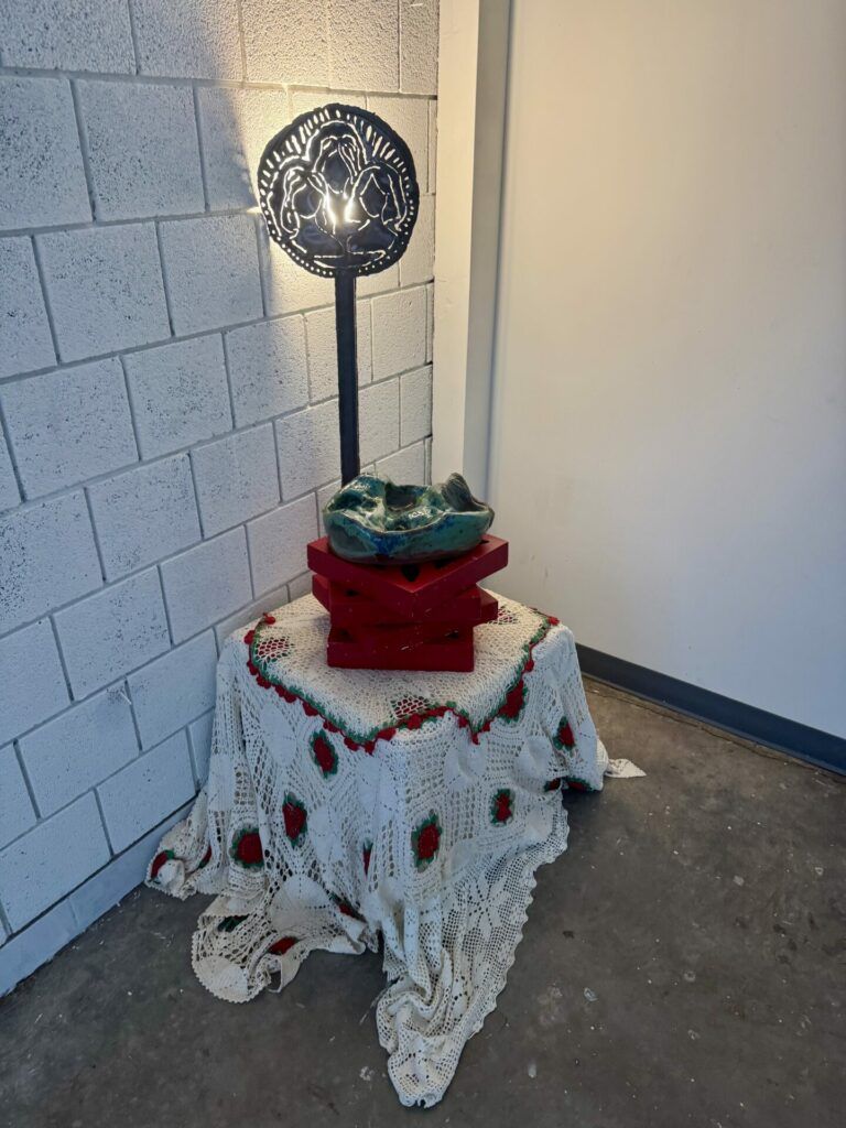 A standing installation that includes a large crochet blanket with red breeze blocks stacked on top. There is a ceramic piece on the blocks. Behind these objects, there is a metal cut-out of three women, illuminated by the light behind them.