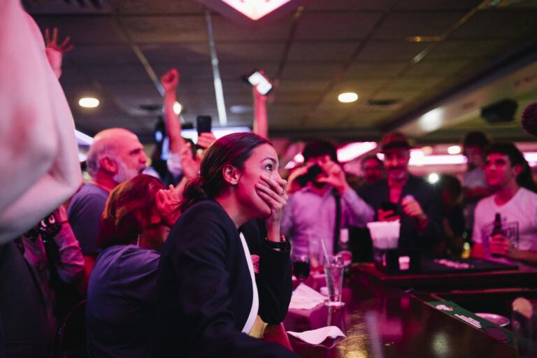 Alexandria Ocasio-Cortez covers her mouth in disbelief in a pink-tinted Park Billiards Cafe.