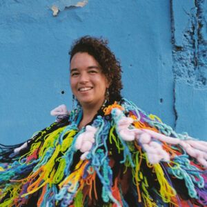 Esotérica Tropical wearing a colorful yarn frock. She stands in front of a blue wall.
