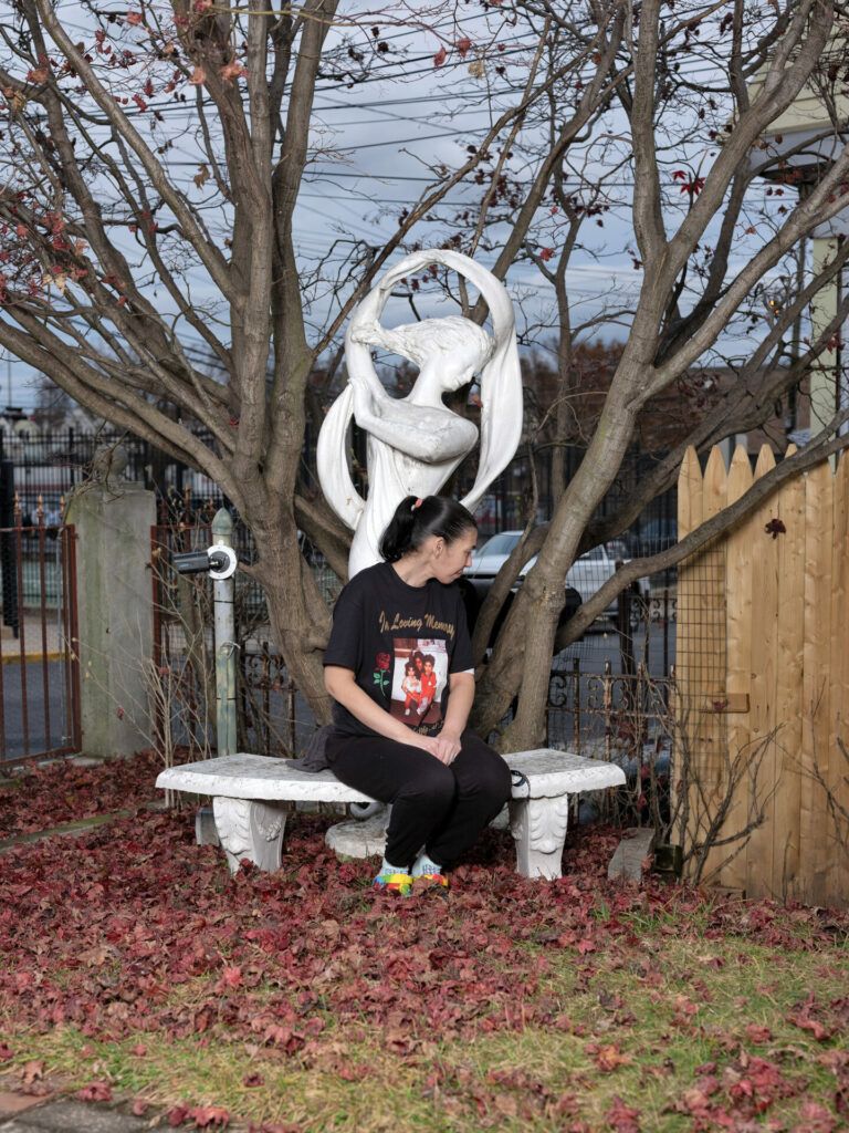 A woman sitting on a marble bench surrounded by red dried leaves. She is wearing a memorial shirt for her son.