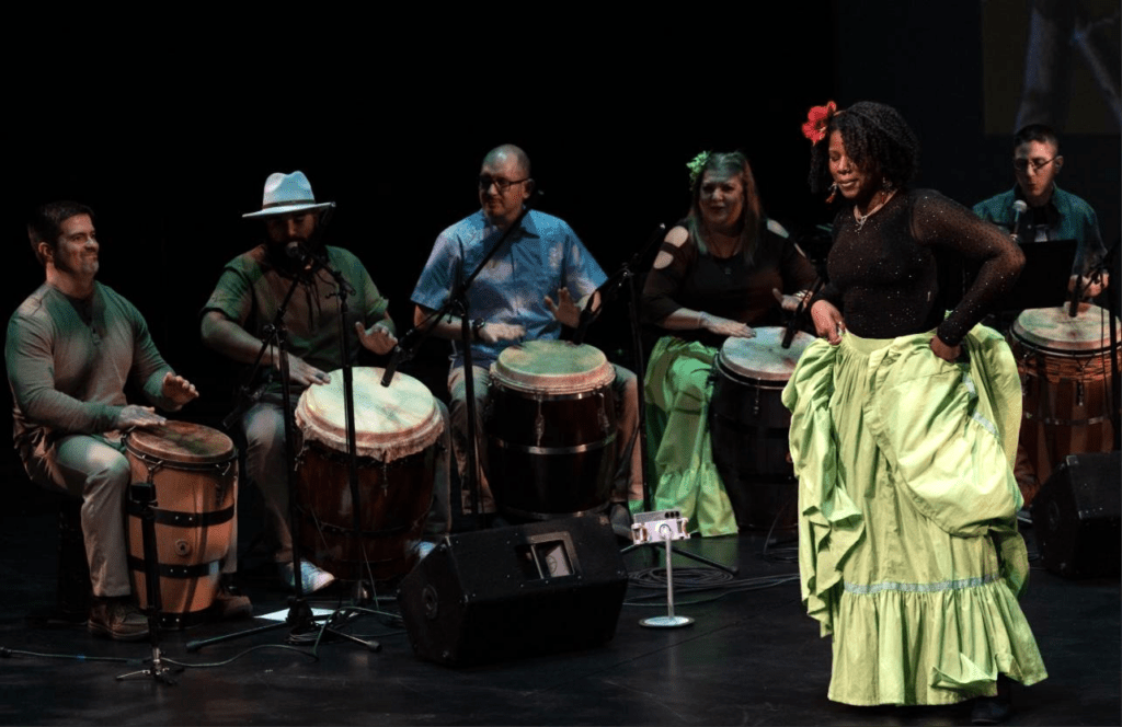 Five people sitting playing bomba music and one dancer in a green traditional skirt.