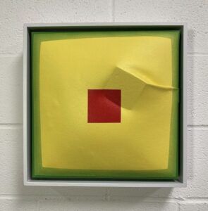 Yellow painting with two green borders. The center of the painting has a red square. Underneath the canvas, a cube pushes to the surface.