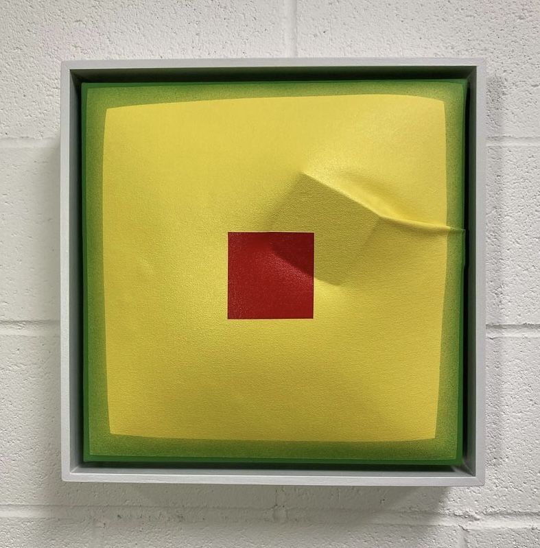 Yellow painting with two green borders. The center of the painting has a red square. Underneath the canvas, a cube pushes to the surface.