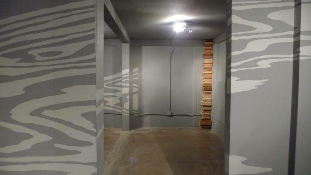 A grey hallway of a basement with a bright white light on the ceiling. The walls are panted in a light grey wave pattern. There is a stack of books in the corner.