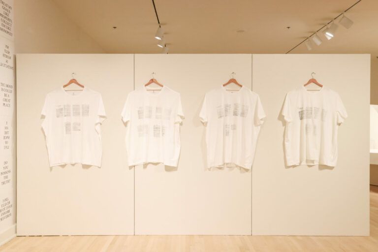 White T-shirts lined up on hangers on the wall. Literature pages are printed onto the shirts in black.