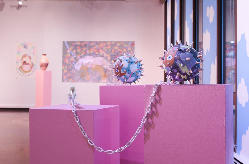 Decorated and painted spiked mace weapons sit on pink display boxes in a gallery.