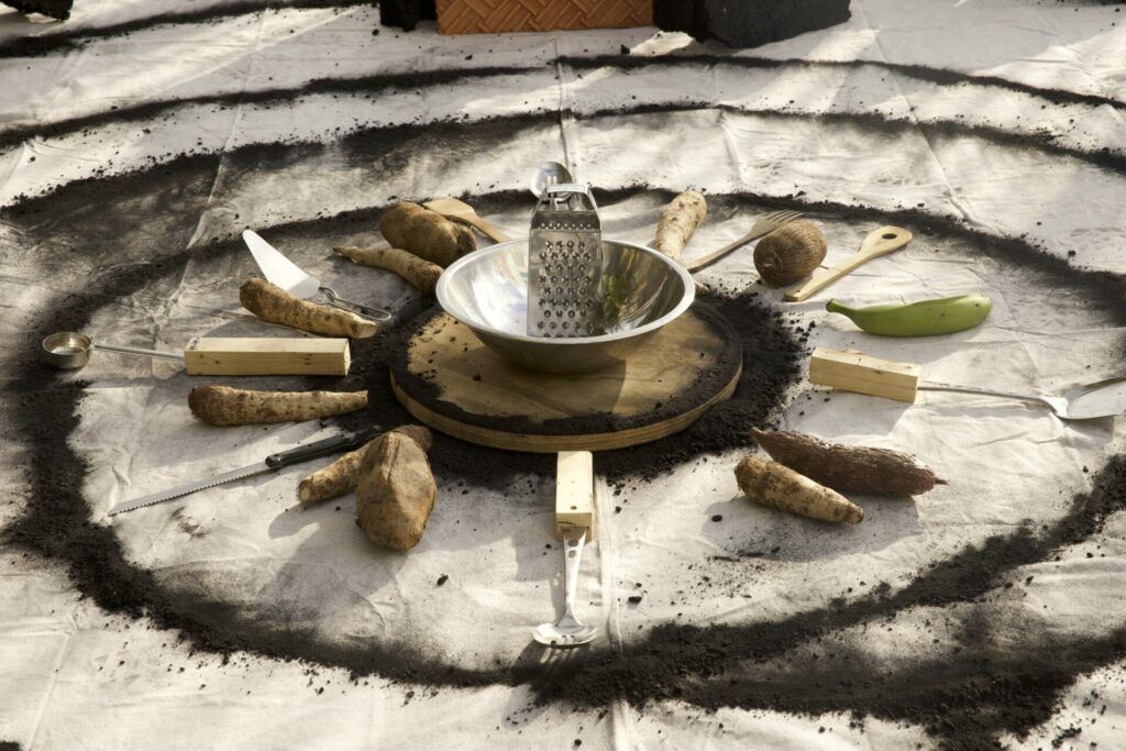 A bowl, grater, and various tools mixed with root vegetables and platanos are set in a radial position in the middle of concentric charcoal circles on canvas.