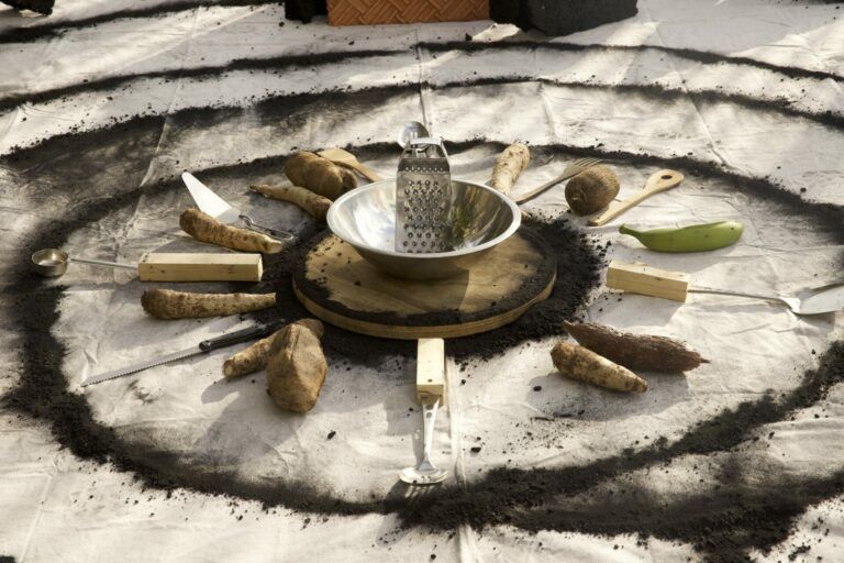 A bowl, grater, and various tools mixed with root vegetables and platanos are set in a radial position in the middle of concentric charcoal circles on canvas.