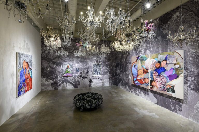 Elaborate installation with chandeliers and wallpaper. Paintings hang on the dark walls.