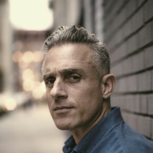 Miguel Luciano against a brick wall looking sternly at the camera.