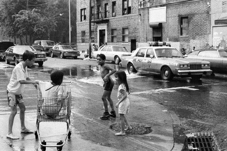 Children playing in an open fire hydrant in the street. There is a police car driving through.