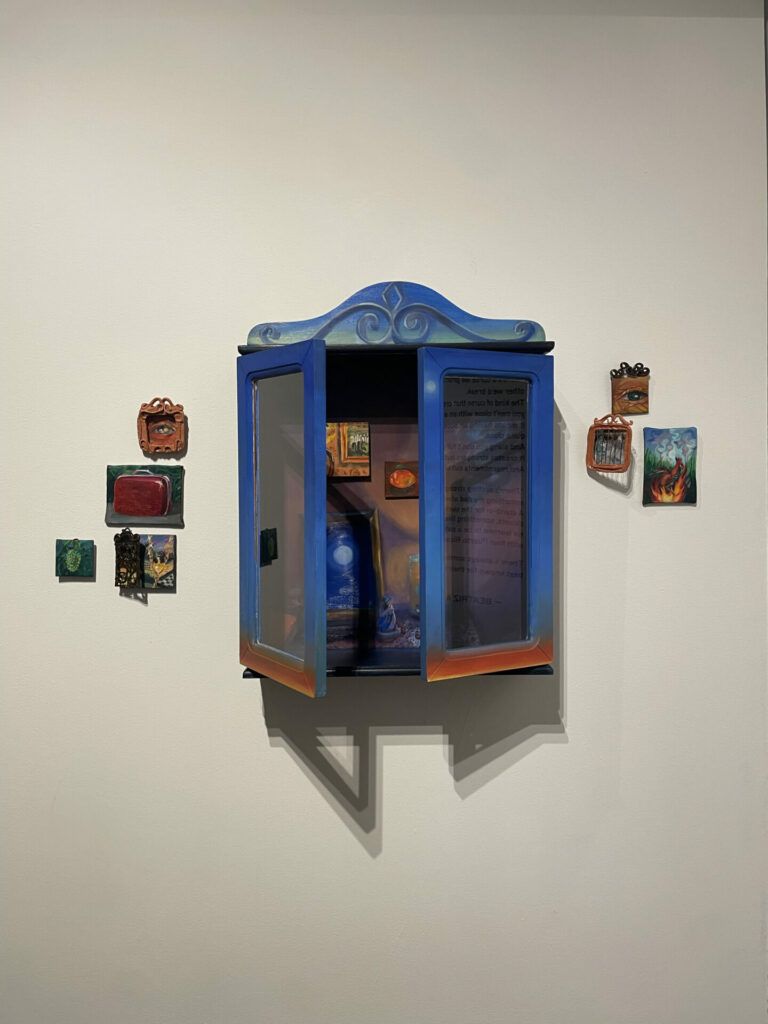 A blue wooden cabinet with paintings and keepsakes inside surrounded by small framed paintings.